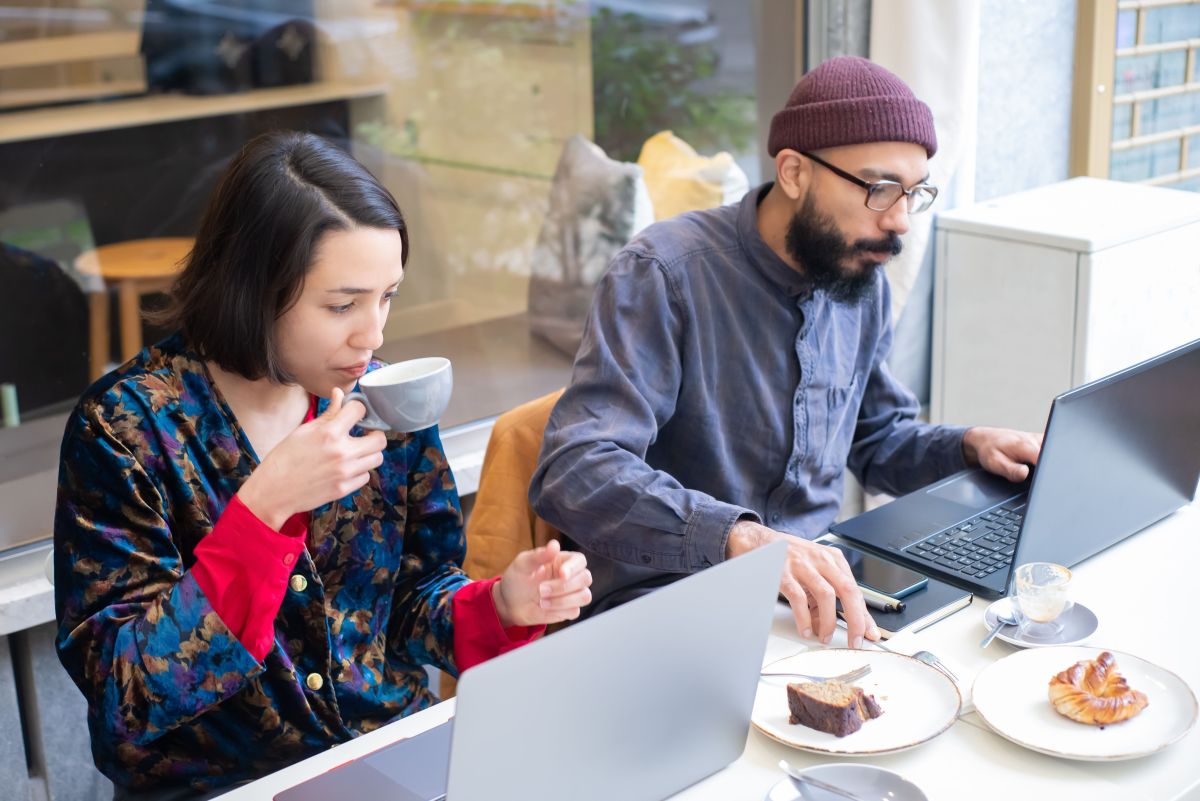 A man and a woman sit outside a cafe with their laptops and coffee.